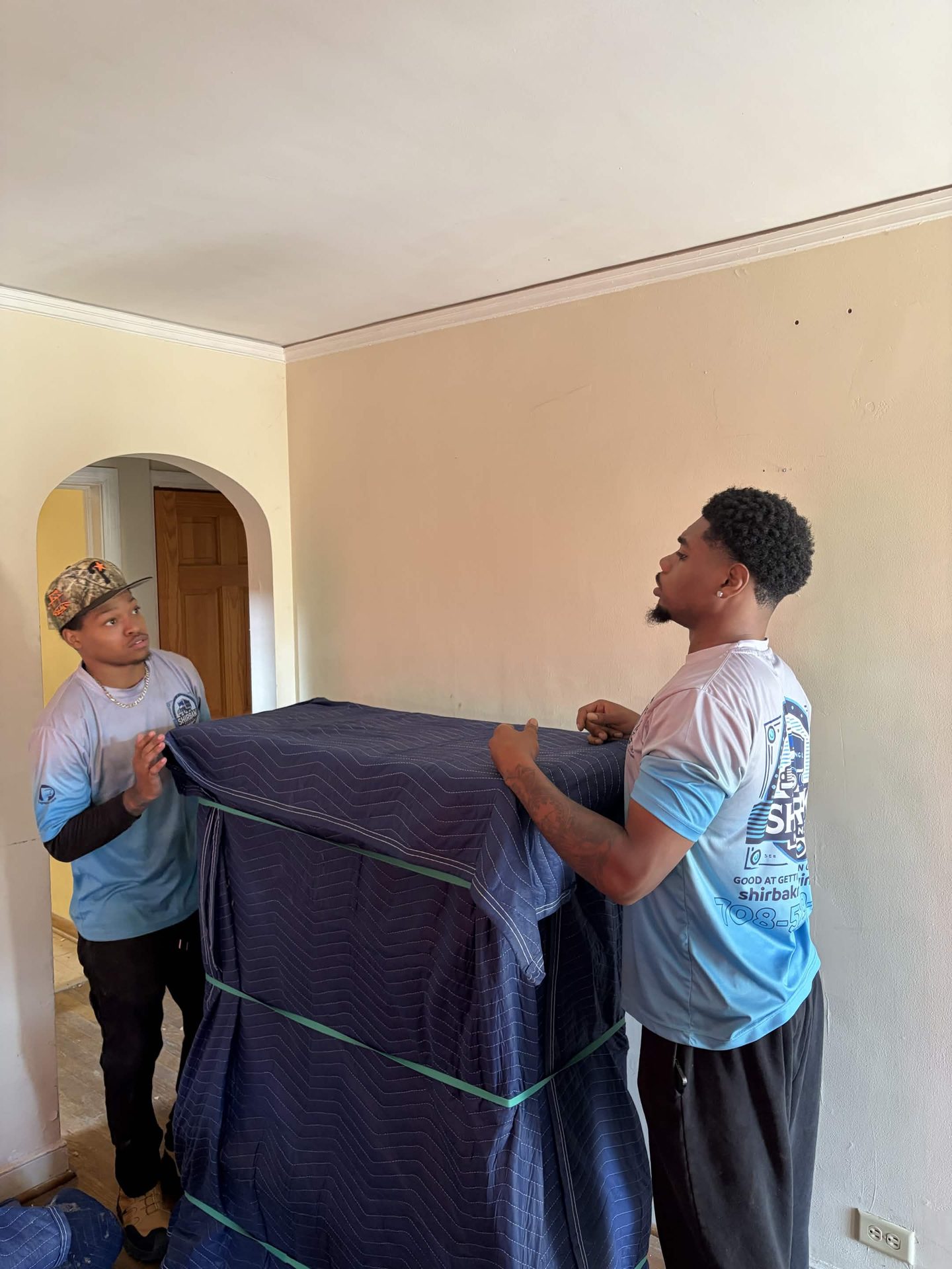 ShirBak movers carry a blanket-wrapped piece of furniture indoors.